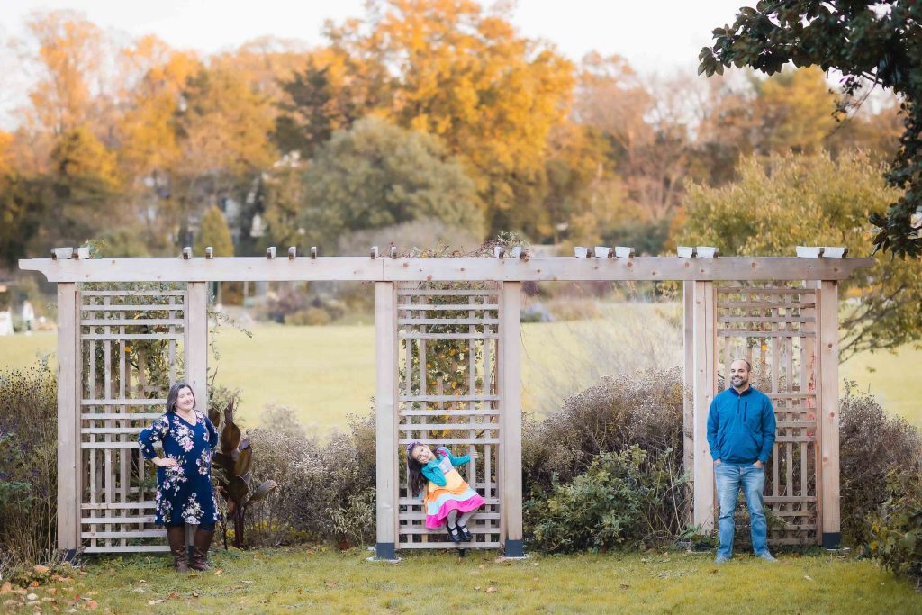 Three people posing outdoors near a wooden pergola amid a garden in Alexandria, Virginia: a woman on the left, a child sitting in the middle section, and a man on the right. Autumn foliage is visible in the background, capturing a perfect portrait of family unity.