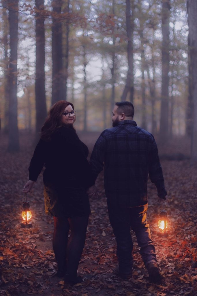 A woman and a man holding lanterns walk together through a forest covered with fallen leaves, creating a moody, almost spooky atmosphere.