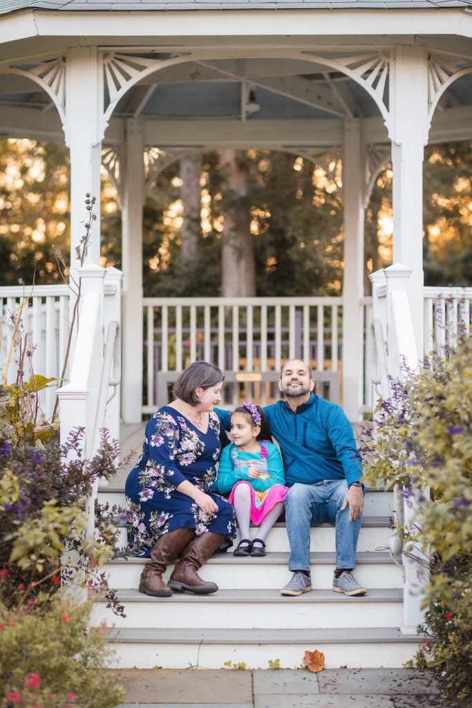 A family portrait captures a serene moment in Alexandria: a woman, a child, and a man sit on the steps of a white gazebo surrounded by foliage. The woman and child hold hands while the man smiles at the camera.