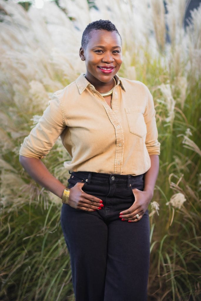 A smiling person stands outdoors with their hands on their hips, wearing a tan buttonup shirt and black pants. They have short hair and are standing in front of tall grasses, creating a perfect portrait for the family album.