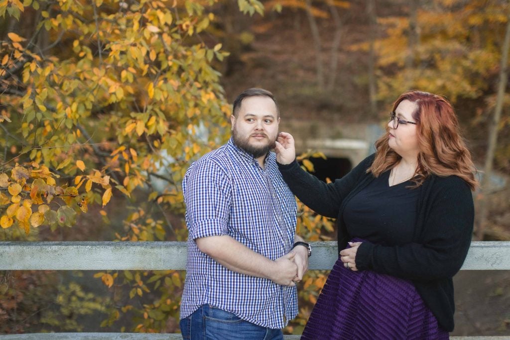 A man in a checkered shirt and a woman in a black top and purple skirt stand on a bridge at dusk, with autumn foliage in the background. The woman touches the man's face.