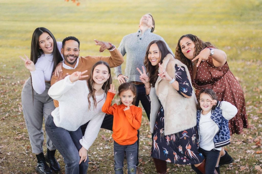 A group of eight people, including adults and children, pose outdoors on the grass at Belair Mansion. Many are making peace signs and playful expressions in this lively family portrait. One person in the back is looking up with their mouth open.