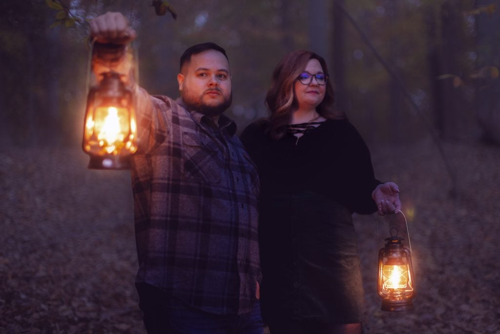 Two people stand in a dimly lit forest at dusk, holding lanterns. The man wears a plaid shirt and the woman, in glasses and a dark sweater, gazes ahead. The scene has an eerie, almost spooky quality to it, like a liveaction portrait of mystery.