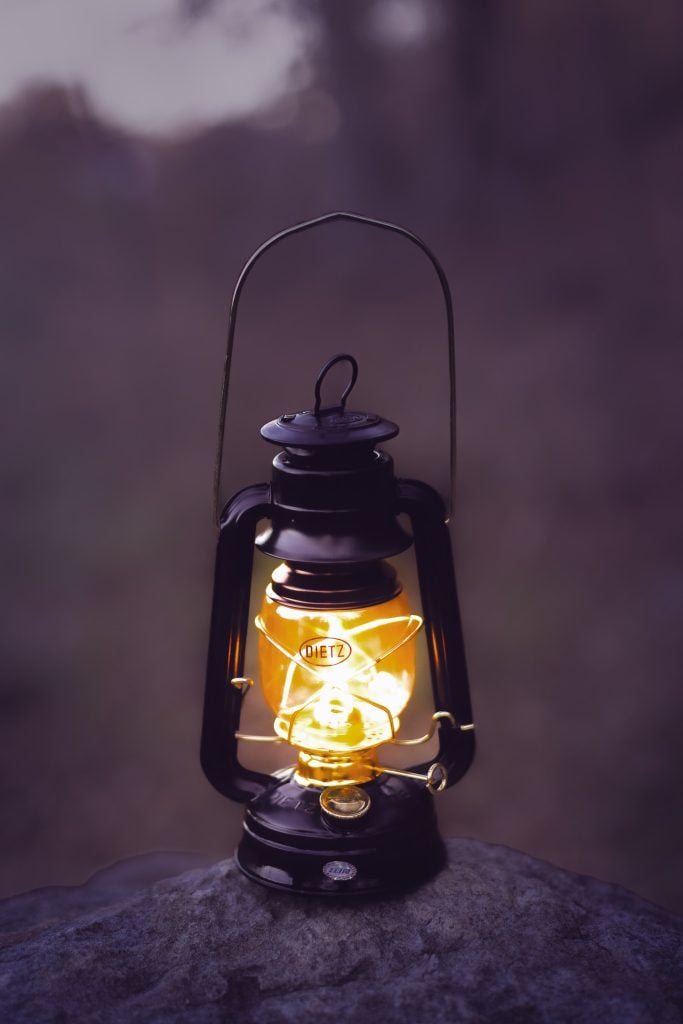 A vintage oil lantern glows with a warm light, placed on a rock outdoors in the dusky evening, creating an almost spooky ambiance.