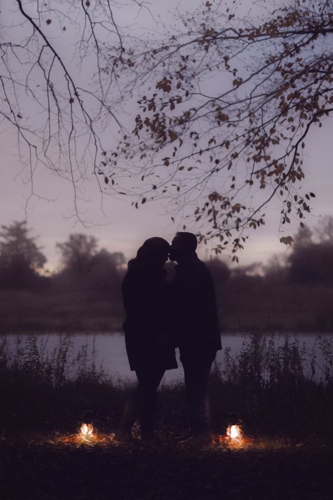 Silhouetted couple standing close together under a tree at dusk, with two lanterns illuminating the ground near their feet. A lake and trees form a serene backdrop, creating the perfect setting for enchanting portraits.