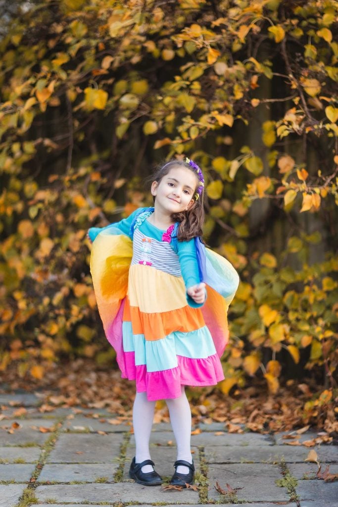 A young girl in a colorful dress and white tights stands on a stone path at Greenspring Garden, surrounded by autumn leaves. She is smiling and holding her arms out, imitating butterfly wings with her dress, creating a perfect family portrait moment.