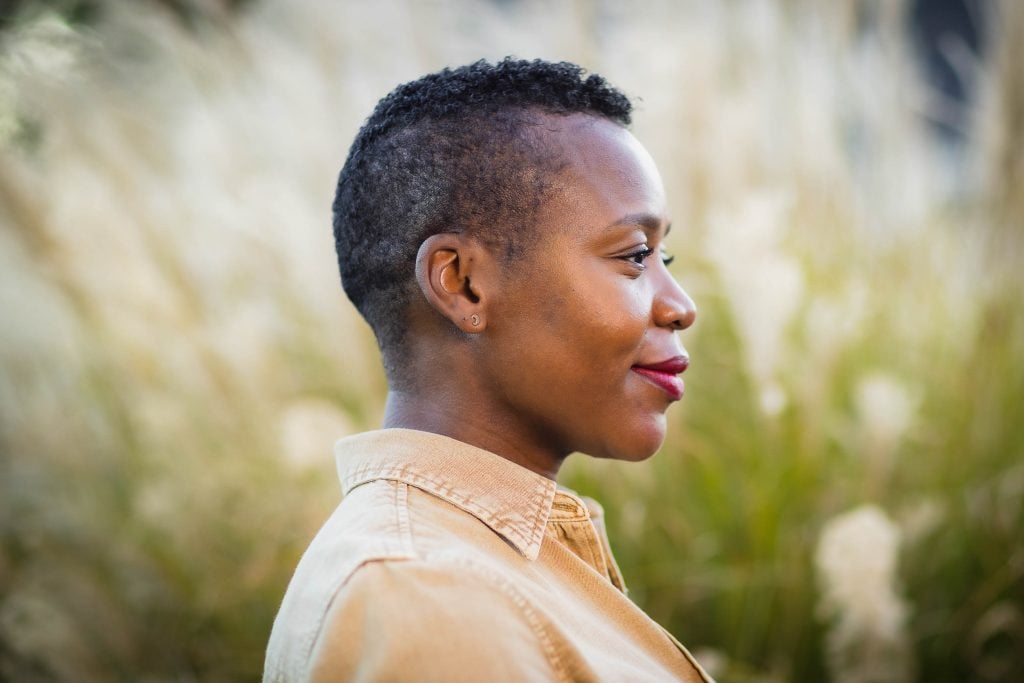 Side profile portrait of a person with short, curly hair wearing a tan collared shirt, smiling slightly with a blurred grassy background.