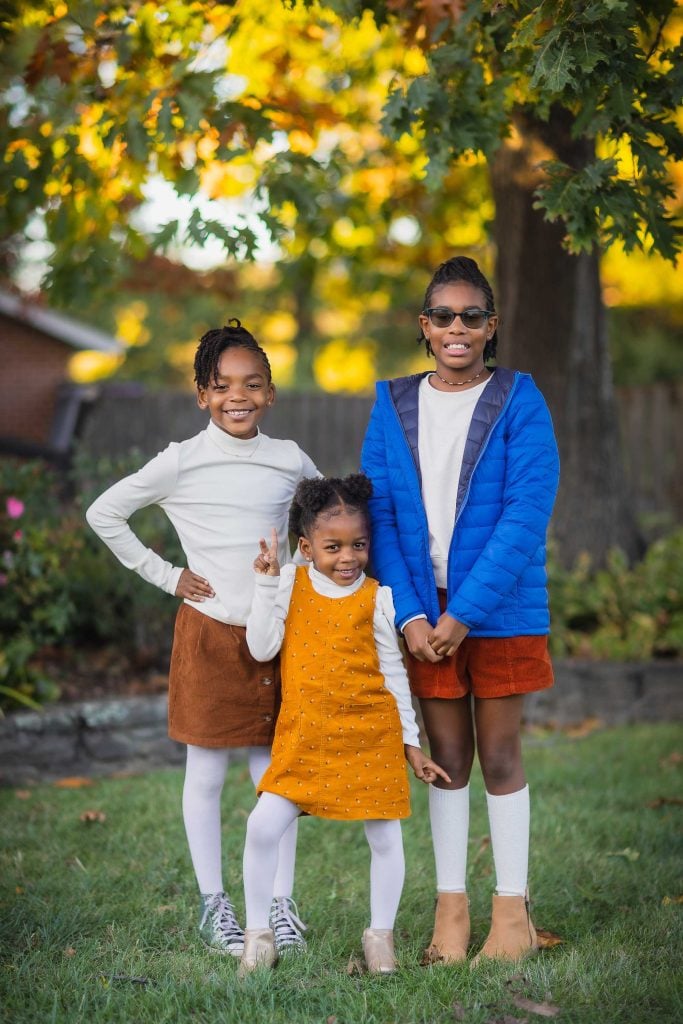 A charming family portrait captures three children standing outdoors with greenery in the background. The child on the left wears a white top and brown skirt, the middle child dons a yellow dress, and the child on the right sports a blue jacket and brown skirt.