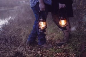 Two people are standing on grass holding lit lanterns in a moody setting. The ground is covered with fallen leaves and dead foliage. One person wears jeans and brown shoes, the other wears a black dress and fishnet stockings, creating a spooky portrait against the eerie backdrop.