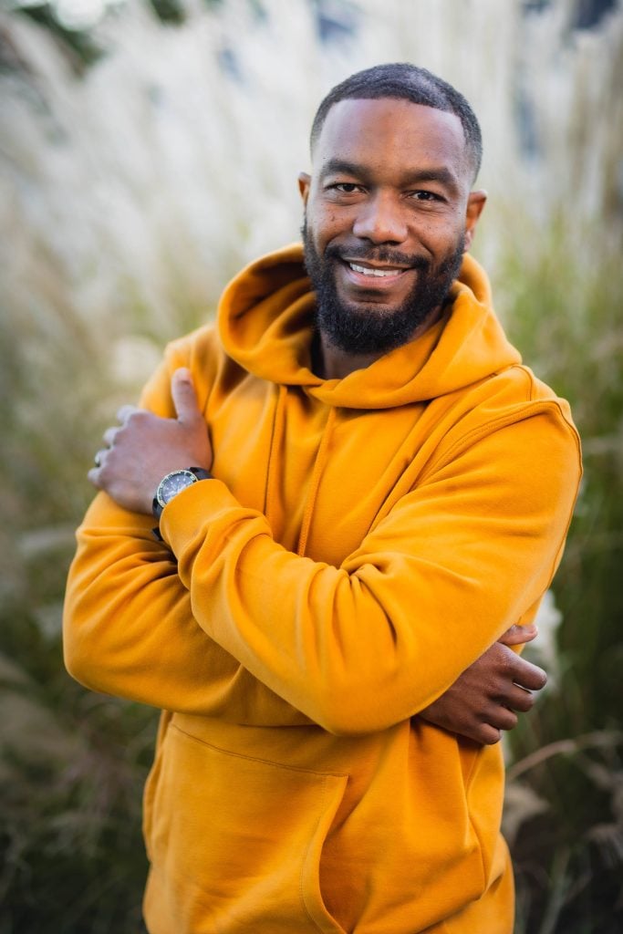 A man with a beard and short hair, wearing a mustard yellow hoodie and a wristwatch, smiles while standing with arms crossed in front of tall grasses, capturing a warm family portrait.