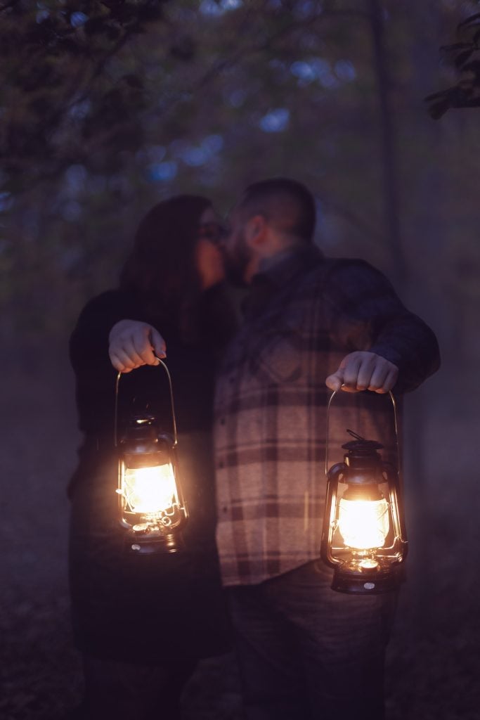 A couple is holding lit lanterns and kissing in a moody, dimly lit forest at dusk.