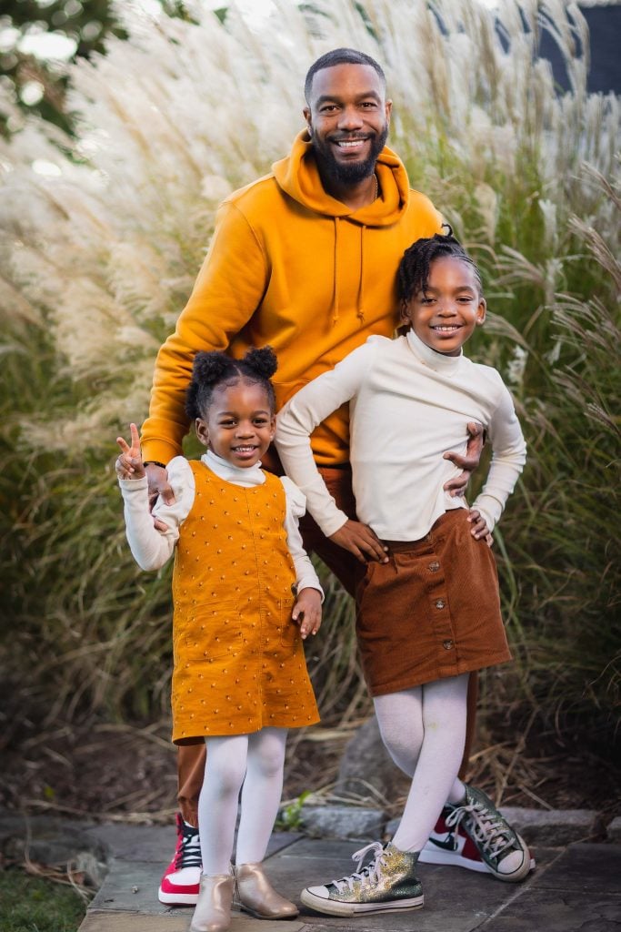 A heartwarming family portrait shows a man and two young girls, all smiling outdoors. The man wears a yellow hoodie, the older girl sports a white top and brown skirt, while the younger girl in a mustard dress with white sleeves makes a peace sign.