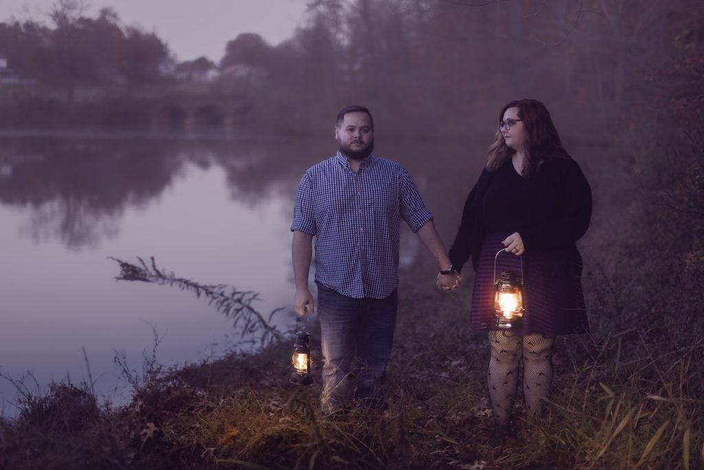 A couple holding lanterns and hands while standing near a calm lake during dusk, surrounded by trees and grass, create a moody scene perfect for portraits.