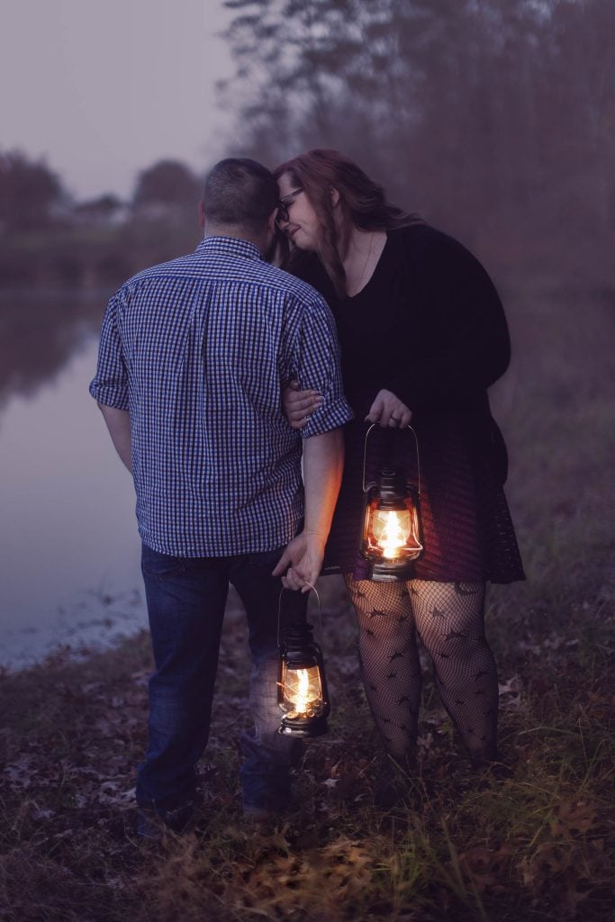 A moody scene unfolds as a man and woman stand by the lakeside holding lanterns. The man faces away while the woman leans in close to him, both enveloped in the spooky dusk landscape.