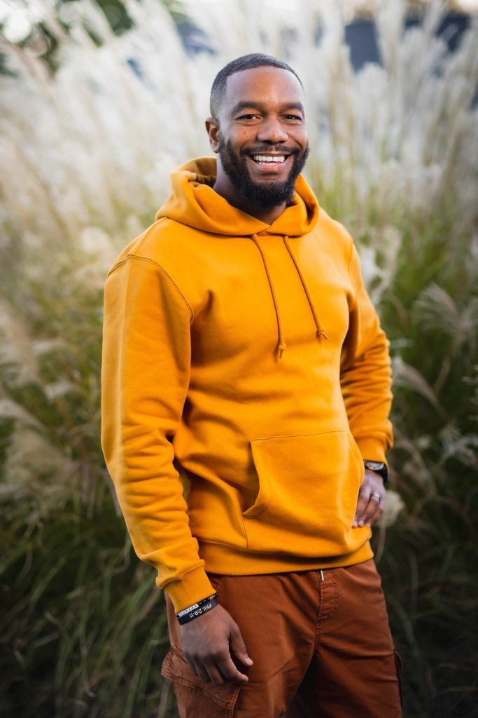 A portrait captures a person wearing an orange hoodie and brown pants, smiling while standing outdoors in front of tall grass.