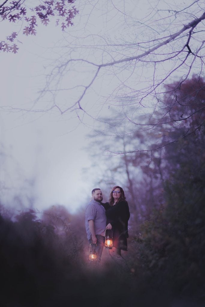 A man and a woman stand outdoors holding lanterns in a dim, wooded area at dusk. The man wears a sweater while the woman dons a dark dress. Bare tree branches loom overhead, adding to the spooky atmosphere of this haunting scene.