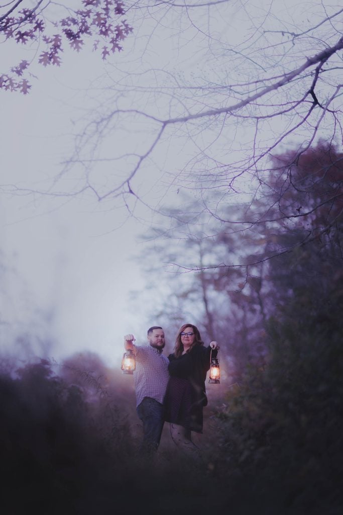 A couple stands outdoors in a misty landscape, holding lanterns under bare tree branches with soft evening light filtering through, creating a spooky yet enchanting dusk scene.