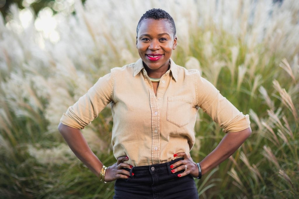 A woman wearing a beige shirt and black pants stands outdoors with hands on hips, smiling at the camera in this charming portrait. Tall grass is blurred in the background.