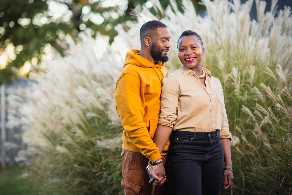 A couple stands holding hands in front of tall, wispy grasses, creating a heartwarming portrait. The man wears a yellow hoodie and the woman a beige shirt. Both are smiling, capturing a moment of simple joy and togetherness.