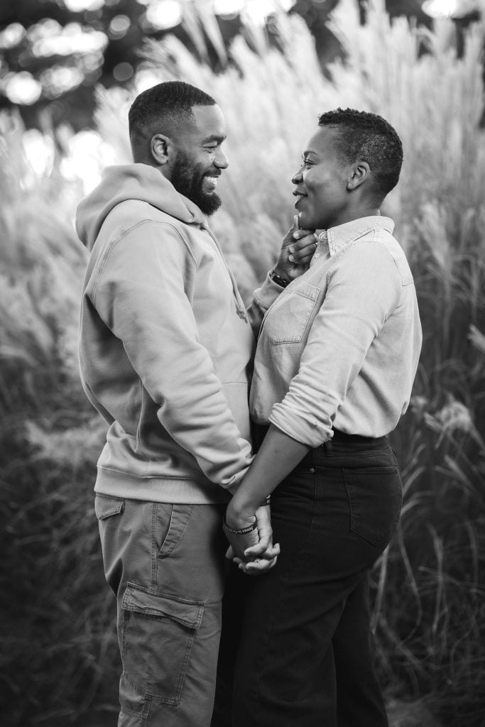 A couple stands outdoors in front of tall grass, holding hands and smiling at each other. In this charming portrait, the man wears a hoodie and cargo pants, while the woman sports a buttonup shirt and jeans.