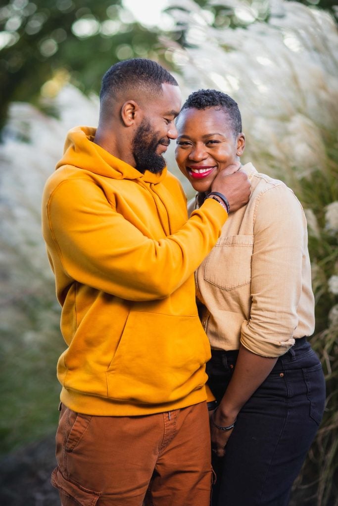 A portrait of a man in a yellow hoodie smiling as he tenderly touches the face of a woman in a beige shirt, who is also smiling. They are outdoors near tall grass.