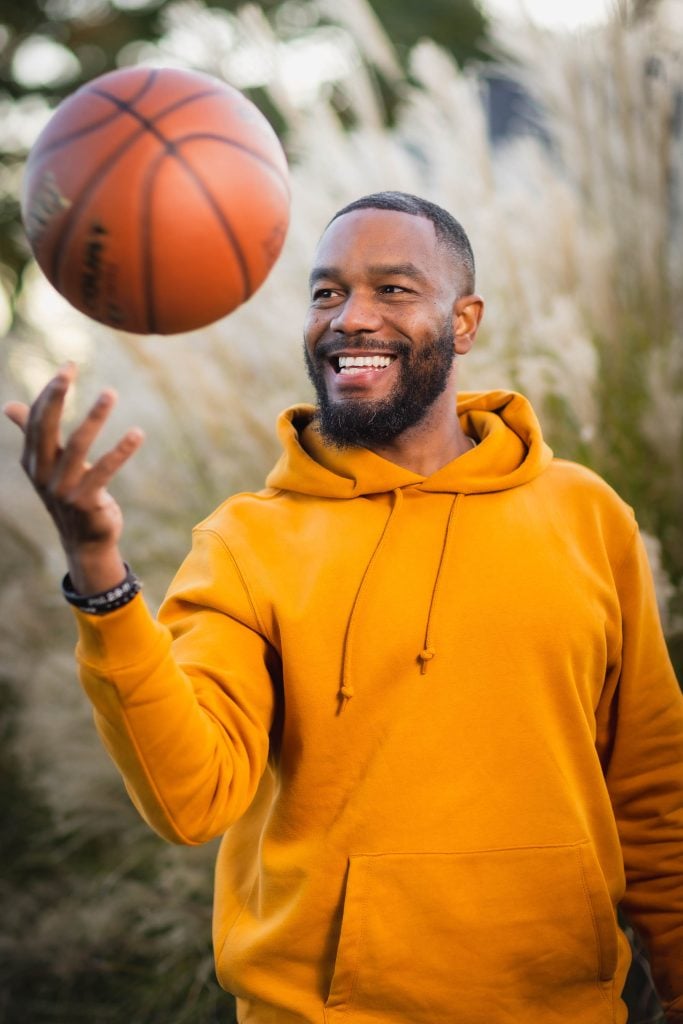 A portrait captures a man in a yellow hoodie spinning a basketball on his finger outdoors, smiling brightly.