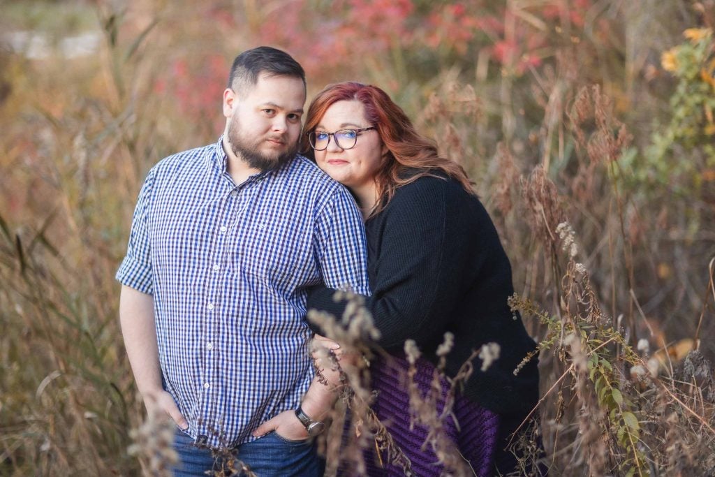 A bearded man in a checkered shirt stands next to a woman with red hair and glasses in a black sweater, leaning on his shoulder, surrounded by tall grass and autumncolored foliage, creating moody portraits that capture the essence of a spooky fall day.