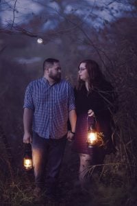 A couple walks through a dimly lit, wooded area at dusk, each holding a lantern. The man's plaid shirt and the woman's skirt and tights add to the scene's rustic charm. Their eyes meet, framing a perfect portrait of evening intimacy with a hint of the spooky in the shadowy surroundings.