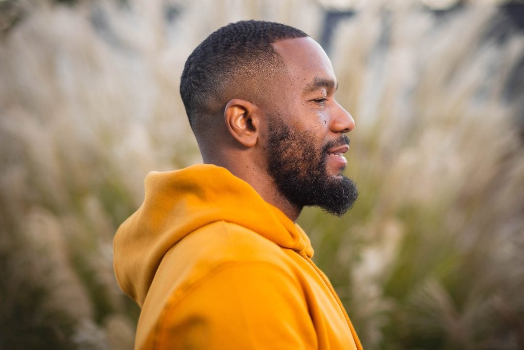 A man in an orange hoodie is standing outdoors, facing to the right. The background is blurry with tall grass, giving the scene a serene portraitlike quality perfect for capturing cherished family moments.