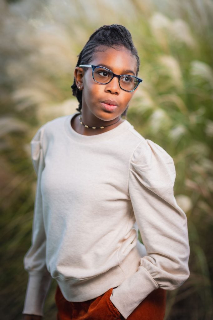 A person wearing glasses and a lightcolored sweater stands outdoors with one hand in their pocket, gazing off to the side. The background features tall grass, capturing a serene family portrait moment.