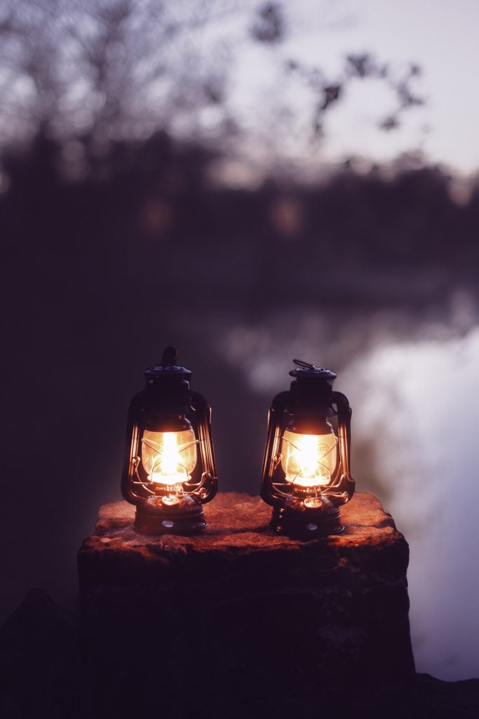 Two glowing lanterns placed on a stone surface near a body of water at dusk, with the moody silhouettes of trees in the background.