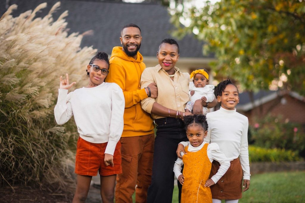 A family of six standing outdoors, posing for a portrait. The group includes two adults and four children, all dressed in autumntoned clothing with a background of grass and trees.