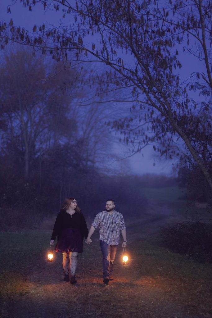 A couple walks hand in hand down a dimly lit path at dusk, each holding a lantern. Trees with sparse branches frame the moody scene, and the sky is a deep twilight blue.