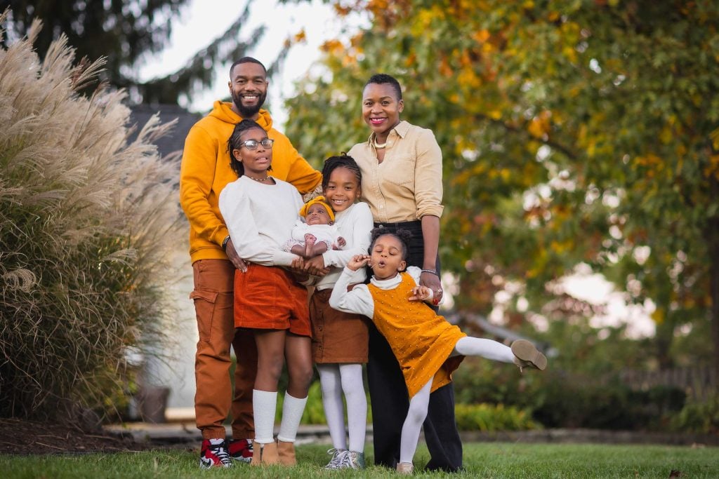 A portrait of a family of five posing for a picture outdoors in front of trees. Two adults stand behind three children, all smiling. One child strikes a playful pose with a leg extended.