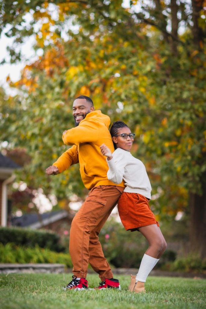 A family portrait features an adult and a child wearing fallcolored clothing, standing backtoback on a lawn. They smile and pose playfully with autumn trees in the background.