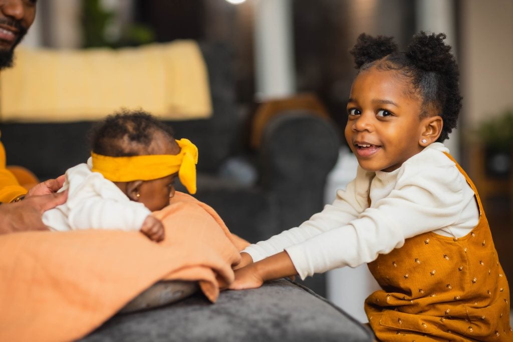 In a charming family portrait, a young child in a white sweater and mustardcolored overalls smiles at the camera while sitting beside a baby lying on a blanket. The baby wears a yellow headband with a bow, completing this heartwarming scene.