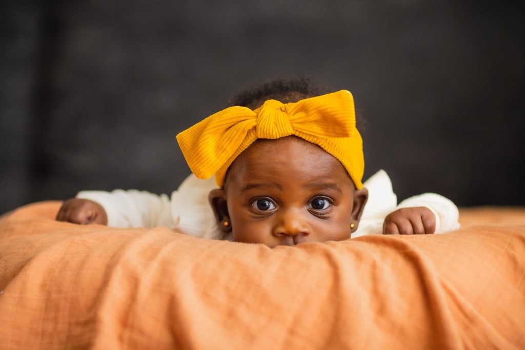 A baby wearing a yellow headband lies on an orange blanket, looking towards the camera with wide eyes, creating a perfect family portrait.