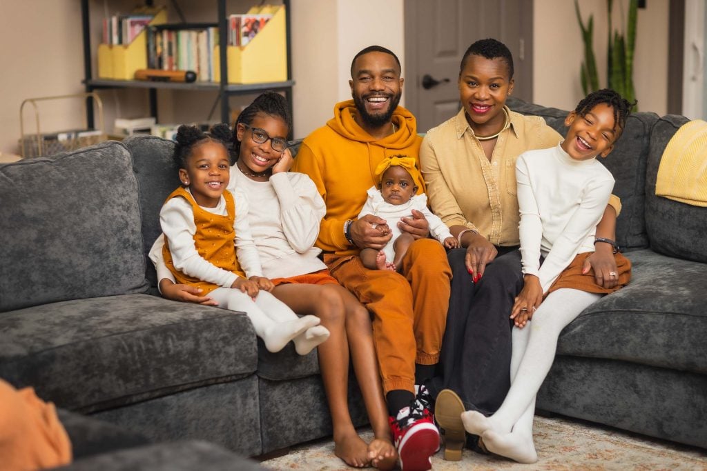 A family of six, including a baby, sits together on a gray couch in a living room, all smiling at the camera for a charming family portrait. The three children wear matching outfits while the two adults are casually dressed.