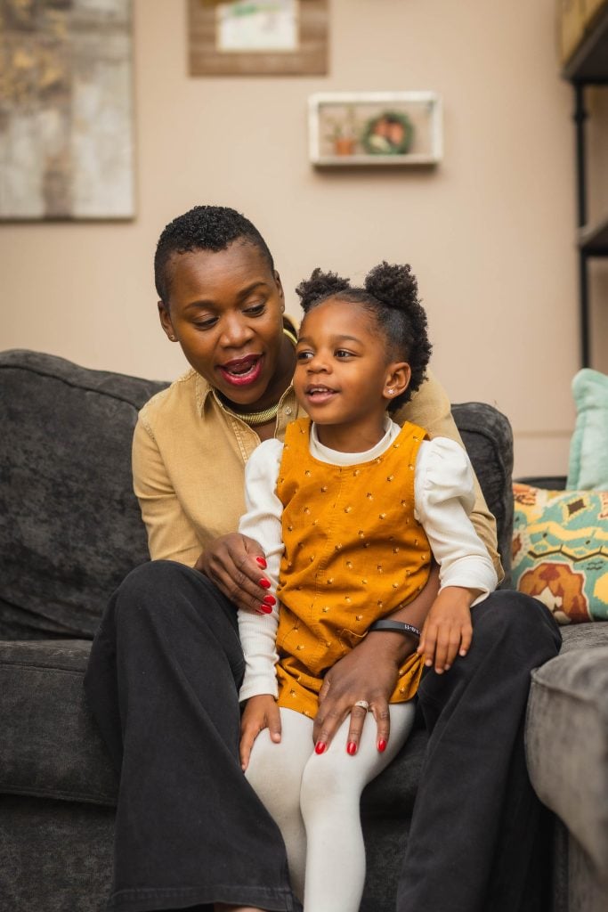 In this heartwarming family portrait, an adult and a child sit together on a couch; the adult holds the smiling child, who is wearing an orange dress over a longsleeve shirt.