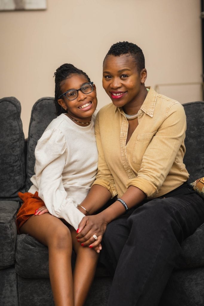 A woman and a young girl are sitting close together on a gray couch, both smiling at the camera in this lovely family portrait. The woman is wearing a beige shirt, while the girl sports glasses, a white shirt, and orange shorts.