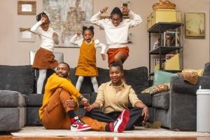 A family of five, with two adults seated on the floor and three children playing energetically behind them on a couch, poses for a charming family portrait in their cozy living room.