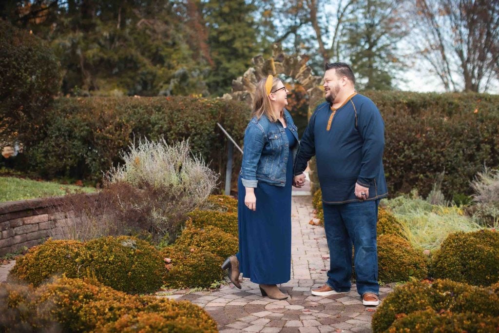 A couple smiling and talking in a garden, surrounded by shrubs and brick pathways, with the woman wearing a denim jacket and blue dress, and the man in a grey hoodie and jeans, perfect for