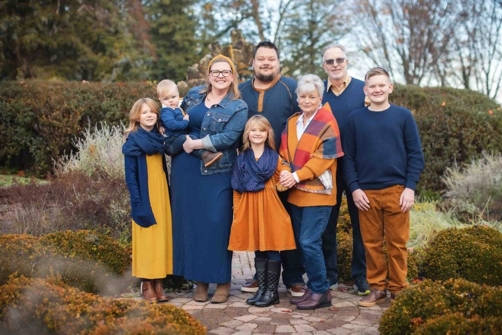 A multigenerational family of eight posing in a lush garden for family portraits, with happy expressions, dressed in casual, colorful attire.