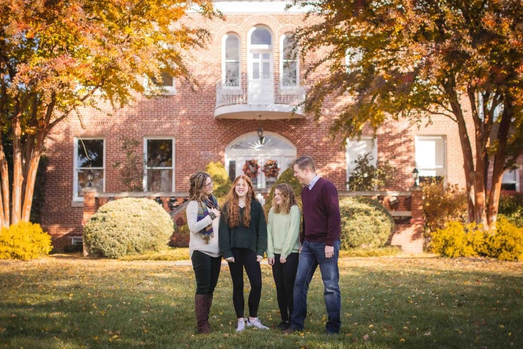 A family of four, two adults and two children, smiling and standing together in a park for family portraits surrounded by autumn trees.