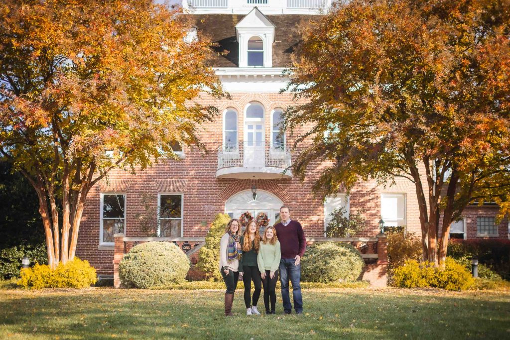 Family portraits of four people posing in front of a brick building with white trim and autumn trees.