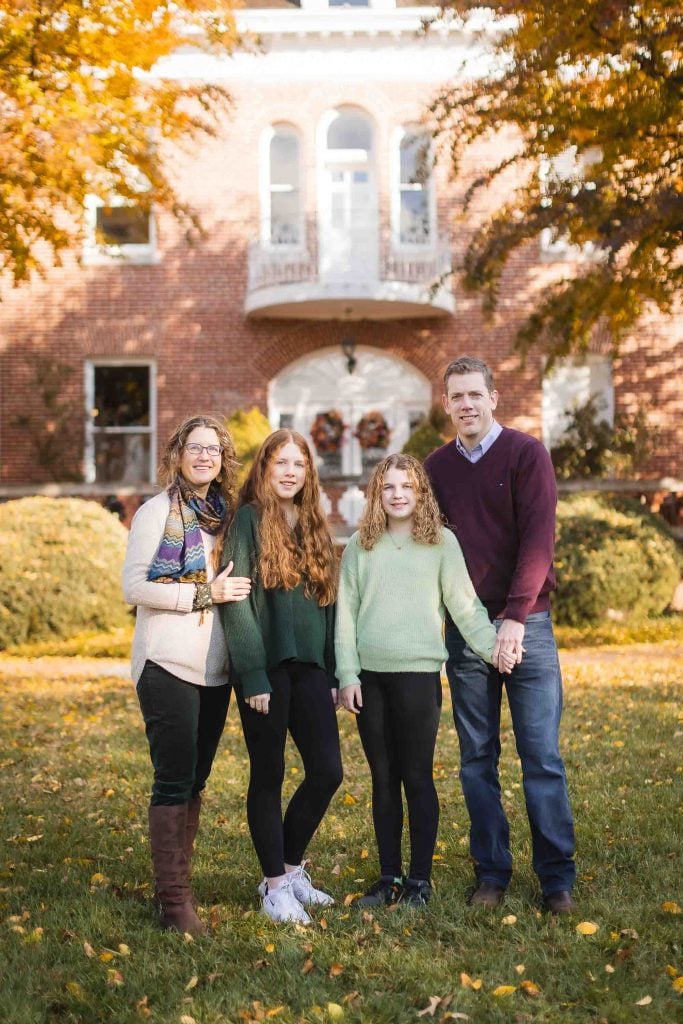 A family of four standing on a lawn in front of a brick house, smiling at the camera during autumn for family portraits.