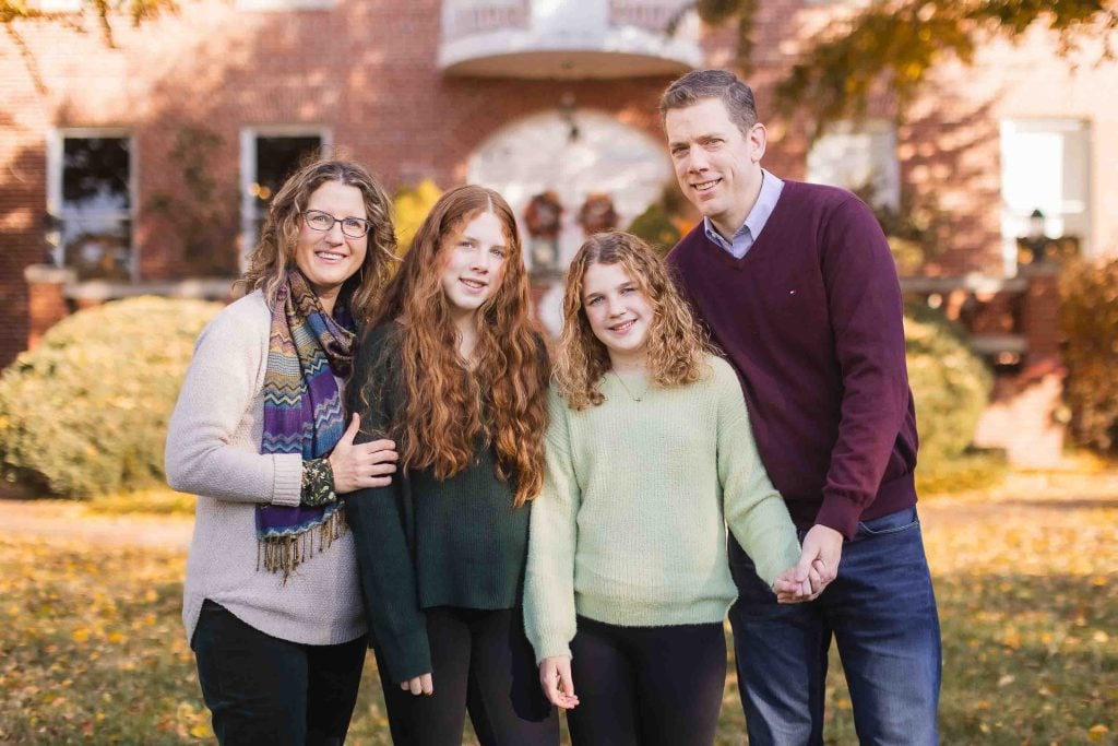 A Family Portraits of four smiling in front of a brick house, featuring two adults and two children, dressed in autumnal attire.