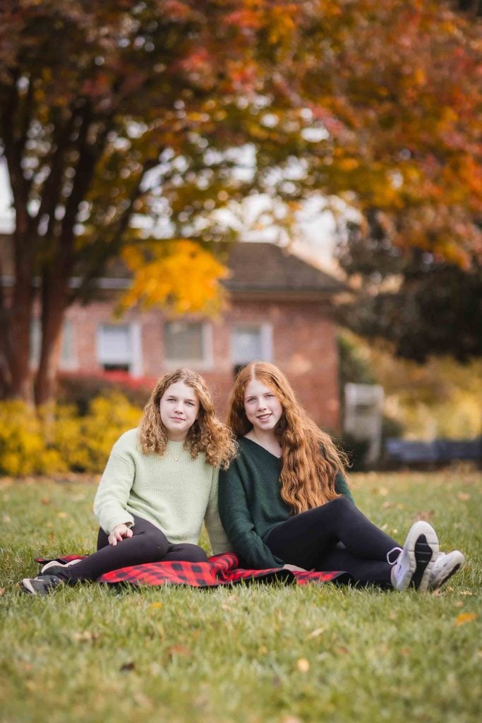 Two young women sitting on the grass with fall foliage in the background, posing for family portraits.