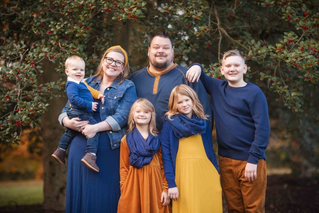 A family of six, with parents and four children, smiling in an outdoor setting with trees and leaves in the background for their Family Portraits.