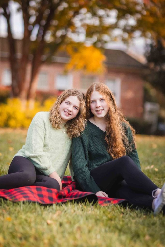 Two young women sitting on a red plaid blanket in a park with autumn leaves around, smiling at the camera for family portraits.